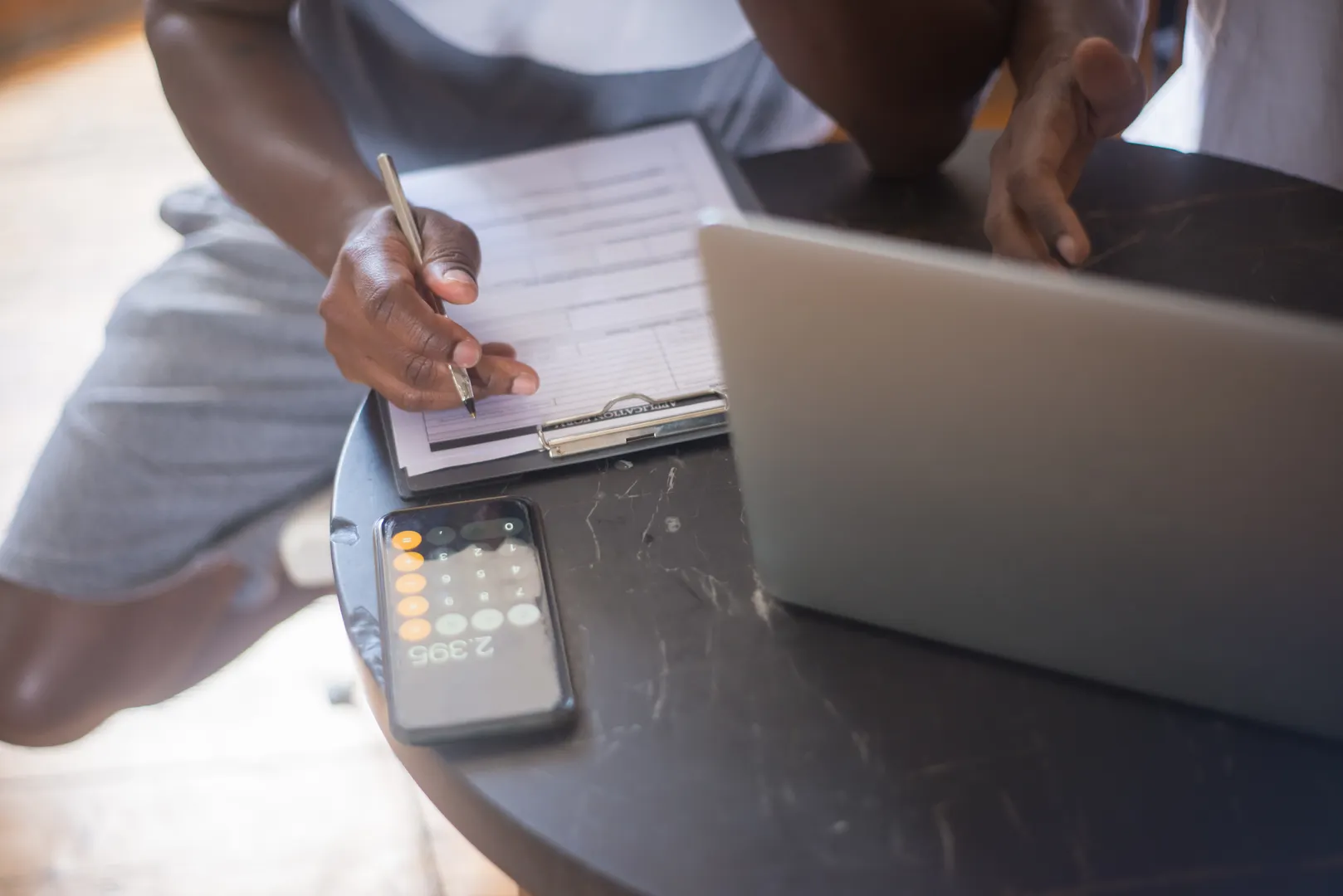 A person working on a laptop with a smartphone and clipboard on a round table.