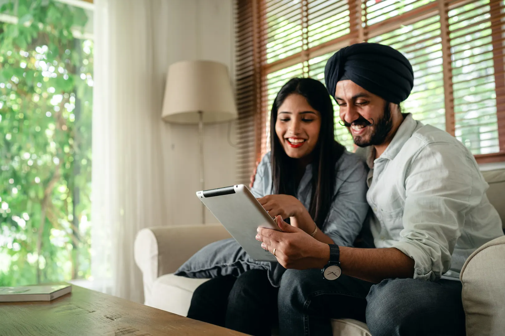 Cheerful Indian man and woman in casual wear sitting on couch in living room near small table and browsing tablet together