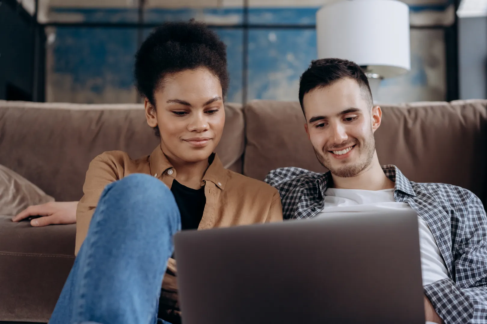 Smiling couple sitting together on a sofa using a laptop, enjoying remote work or leisure time indoors.