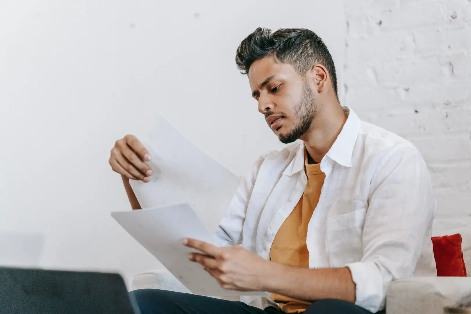 Concentrated ethnic male in casual wear reading documents and sitting on armchair near opened netbook