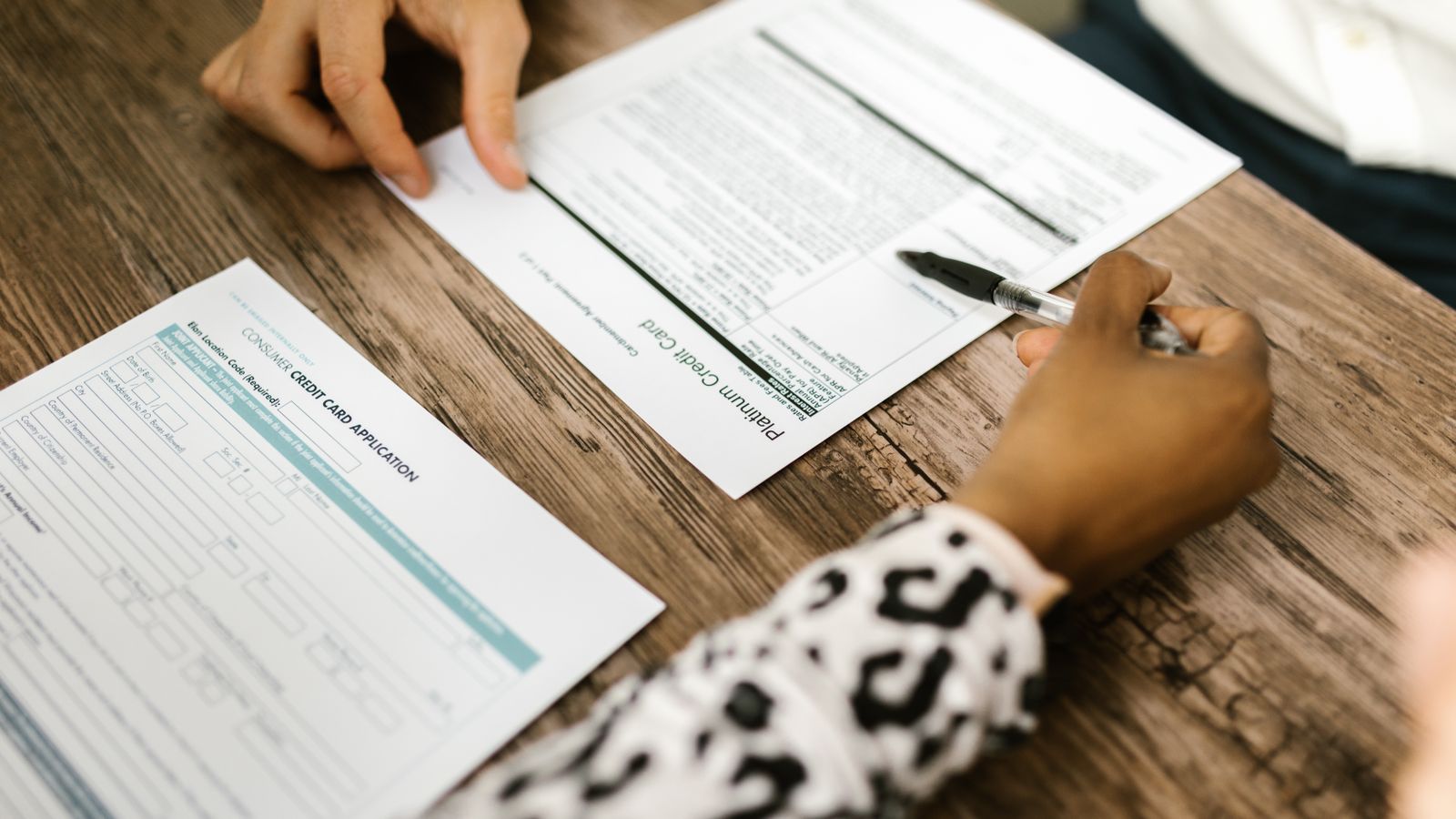 Two people at a wooden kitchen table going through credit card and loan paperwork, one holding a pen over a form