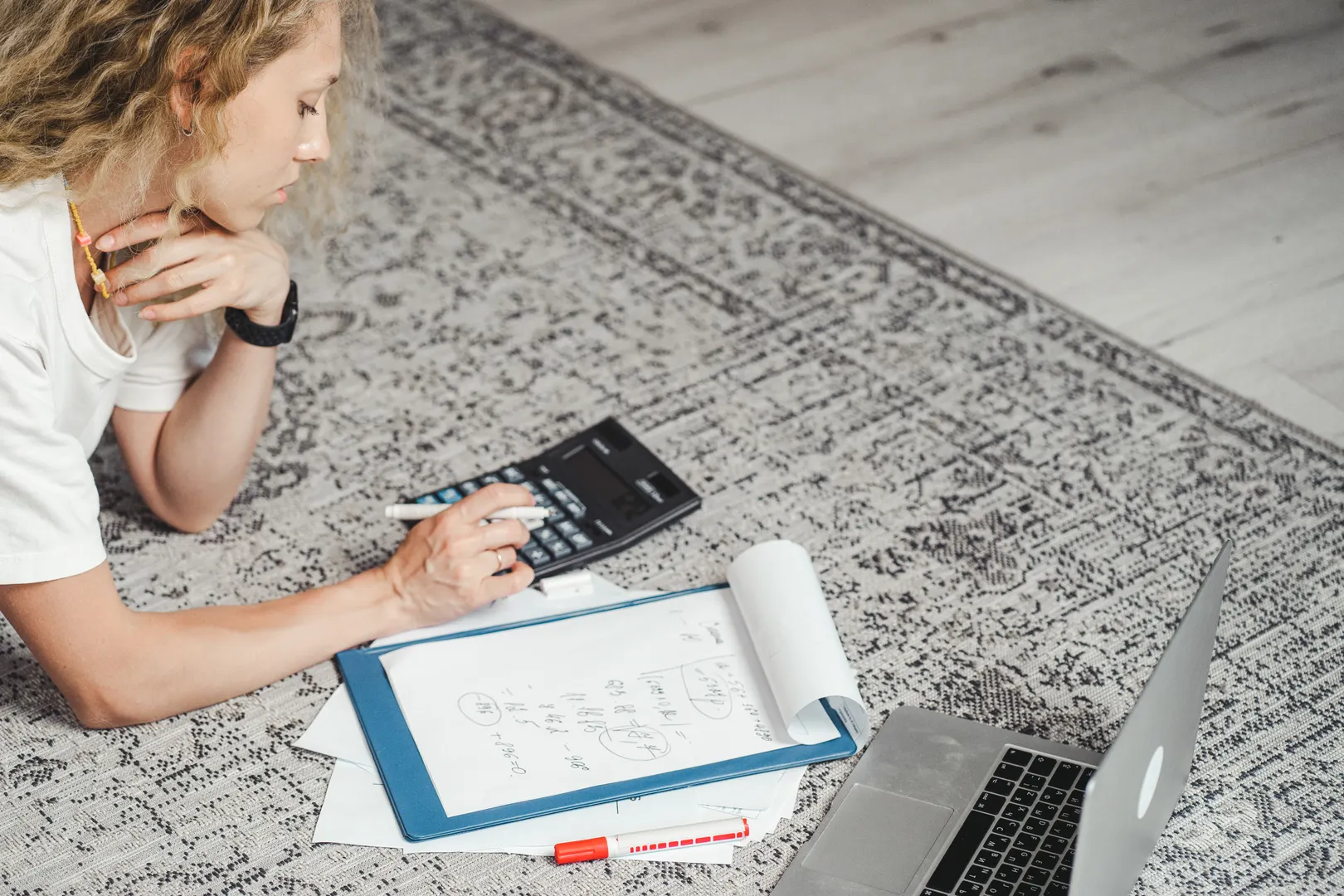 A woman calculating expenses on the floor beside paperwork and a laptop