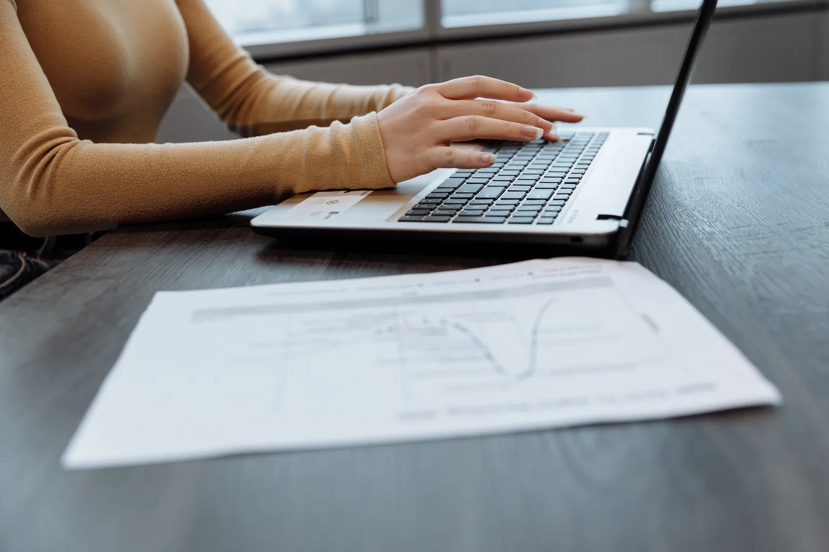 Close-up of hands using a laptop beside a printed financial report
