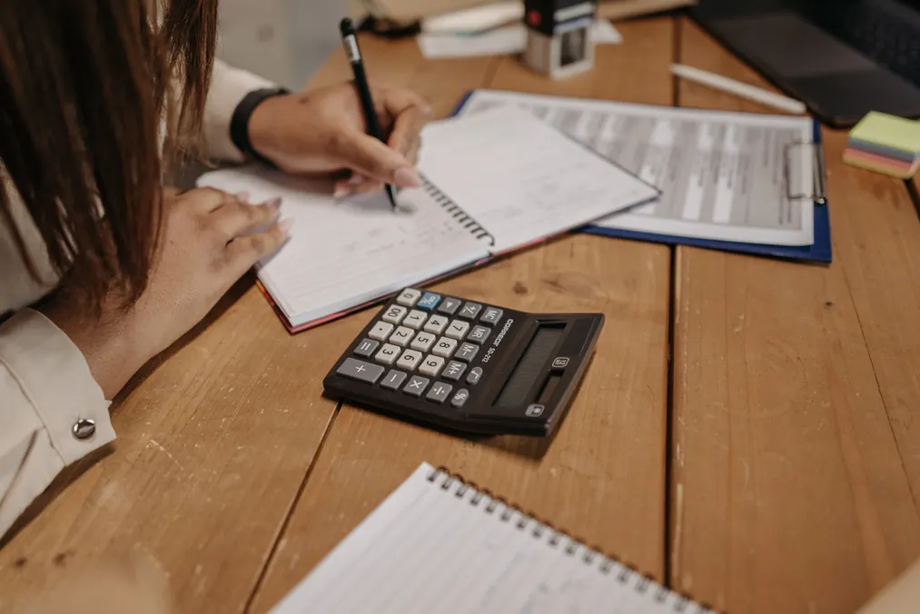 Person writing notes beside paperwork and a calculator