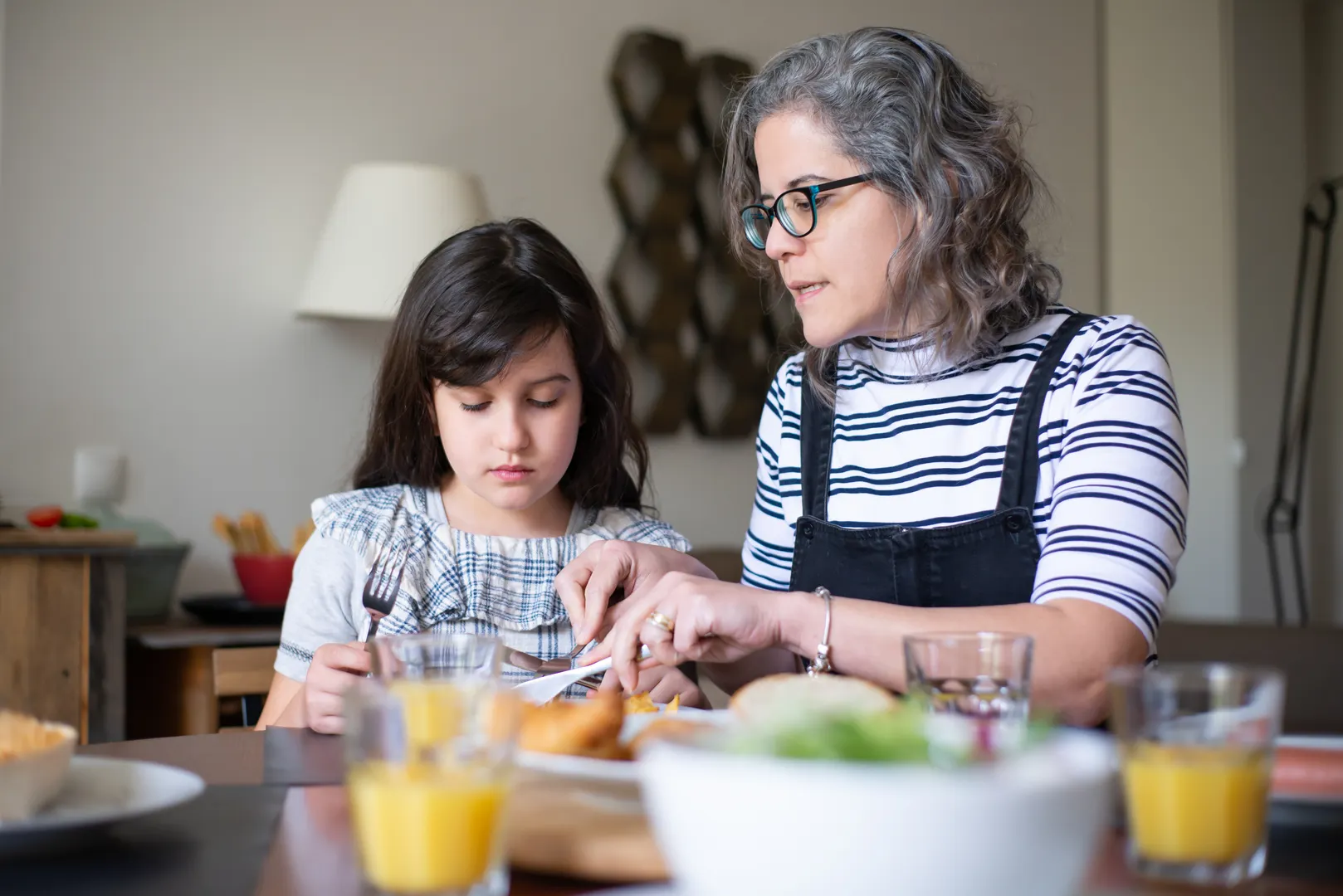 A mother closely helps her young daughter during breakfast at the kitchen table