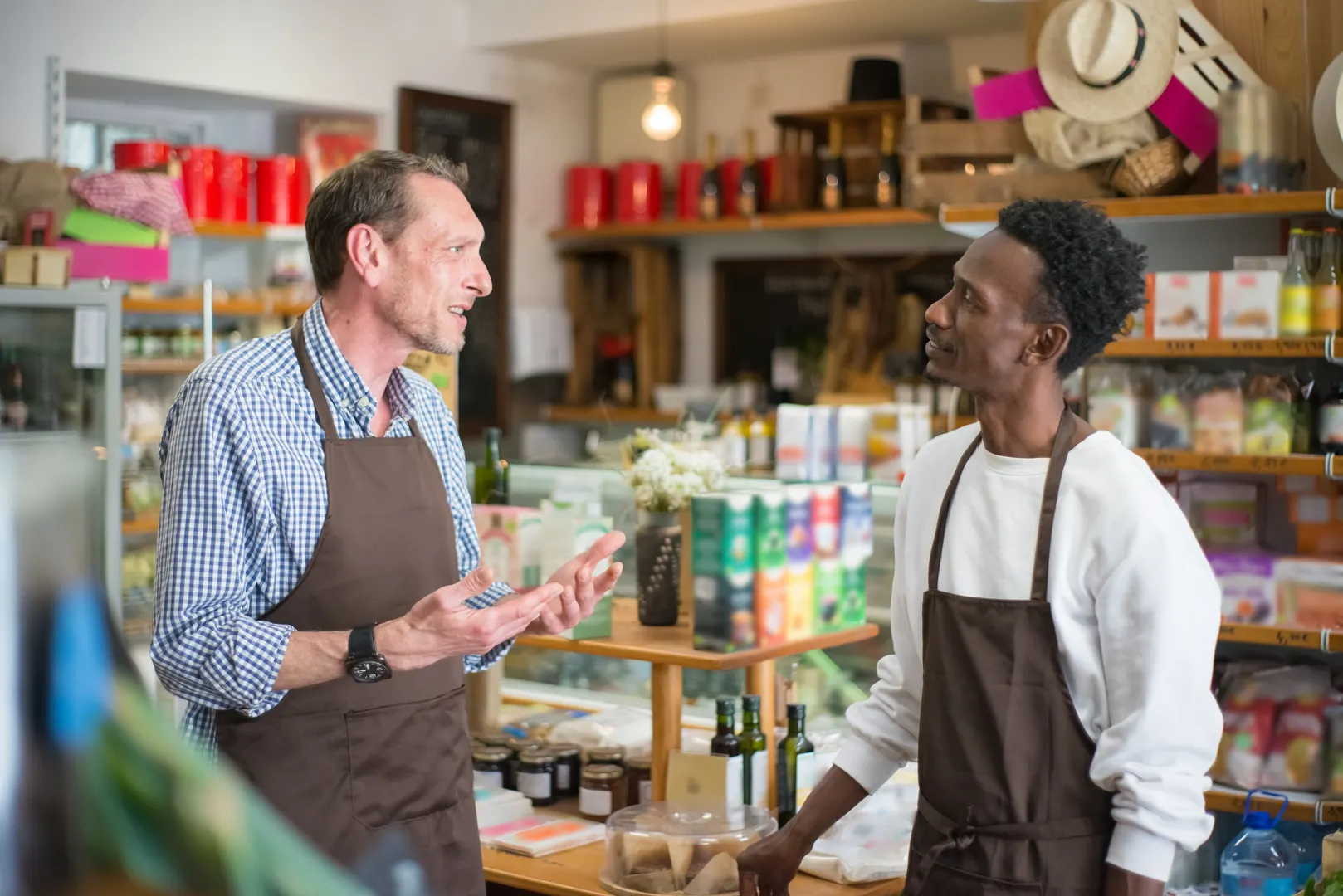 A shop owner talks with a young employee wearing an apron in a small grocery store