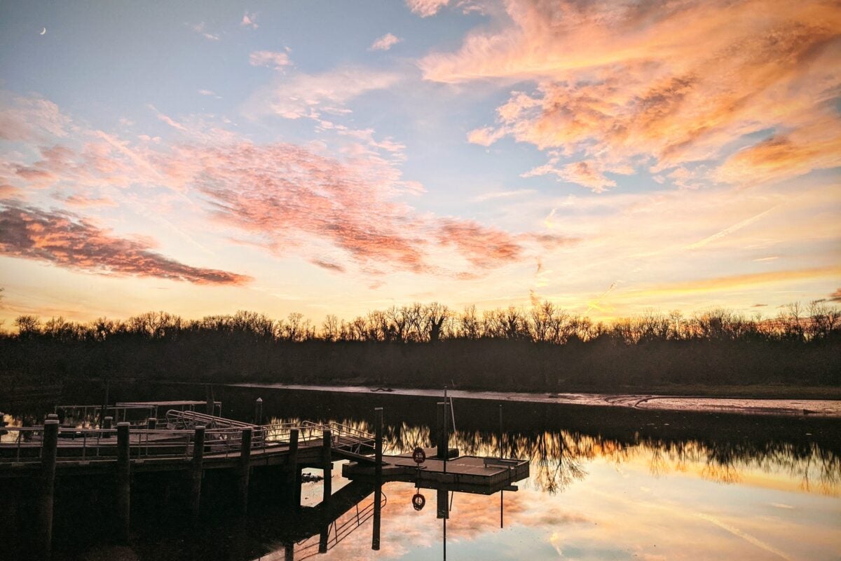 A body of water, a wooden dock, and the far shore beneath a lovely, pink and gold sunset. The water is perfectly calm and reflects the sunset like a mirror.