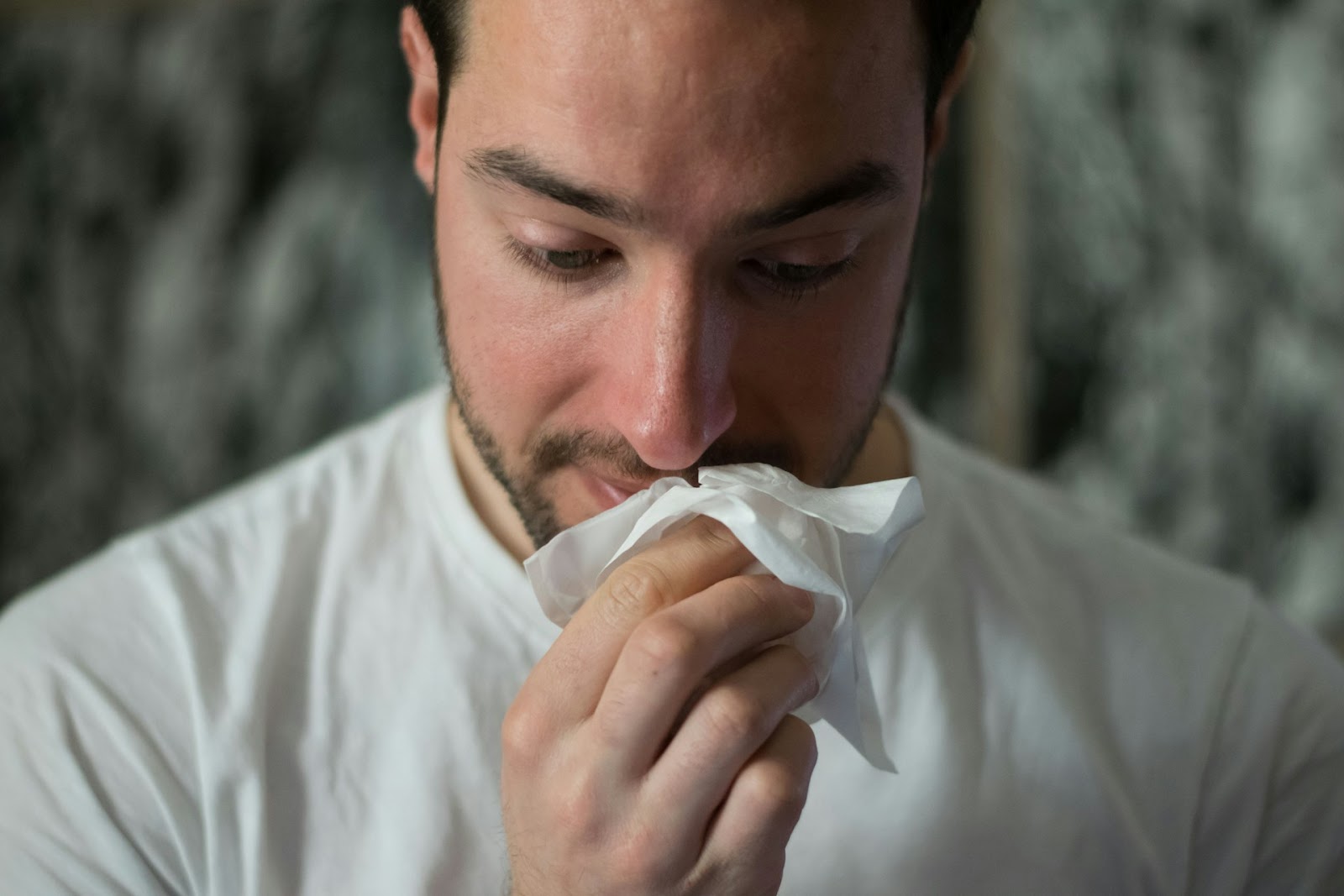 A man is using a tissue to blow his nose, appearing to relieve a cold or allergy symptoms.