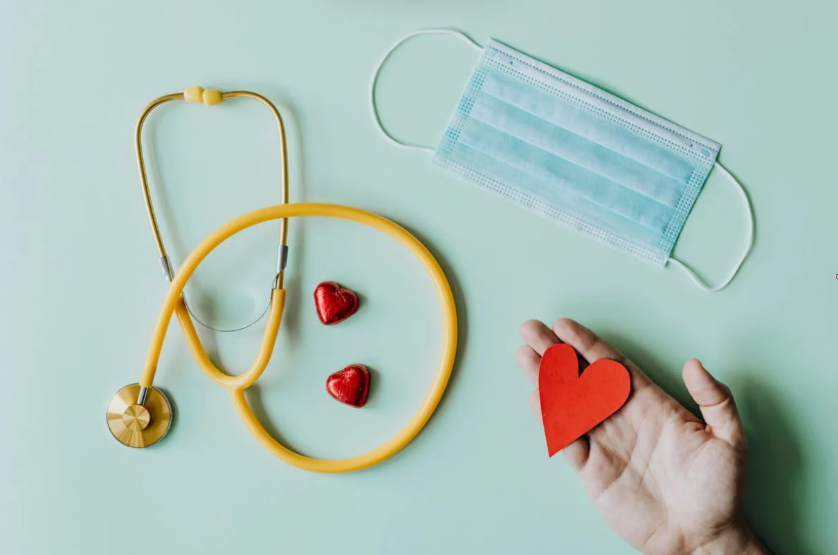 A hand holds a red heart and a stethoscope against a green background, symbolizing care and health.  