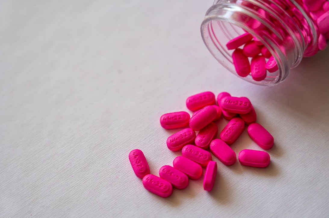 Pink pills spilling from a glass jar onto a white surface, creating a colorful scattered arrangement.