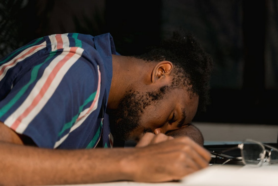 A man dozes off on his laptop, surrounded by work materials, illustrating fatigue from long hours of remote work.