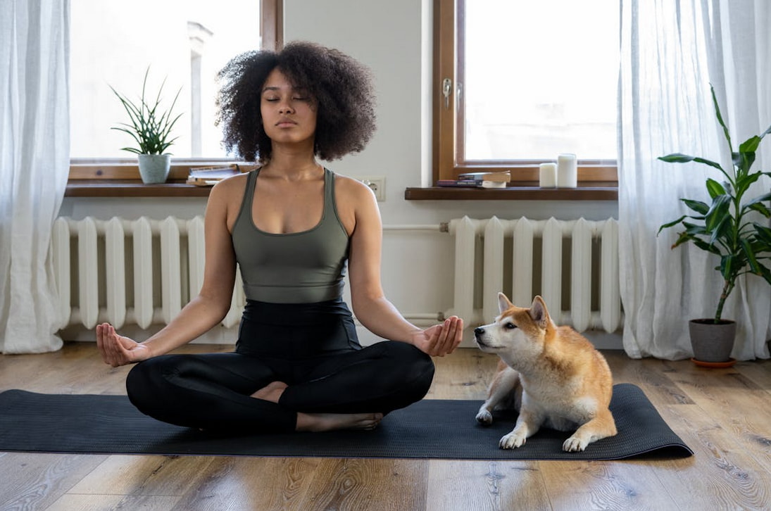 A woman sits cross-legged in meditation with her dog resting beside her, both appearing calm and serene.