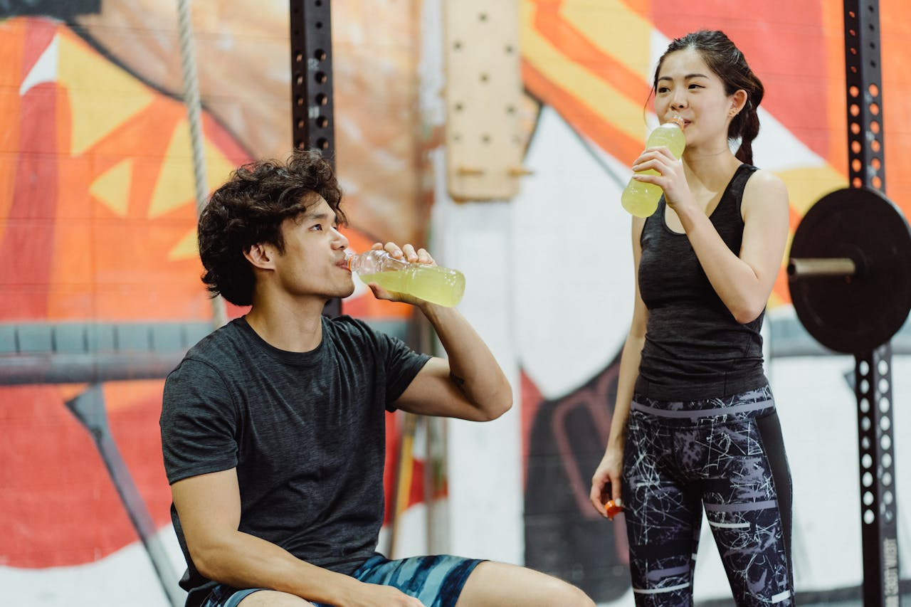 A man and woman are sharing a bottle of water, both taking sips while enjoying a sunny outdoor setting.  