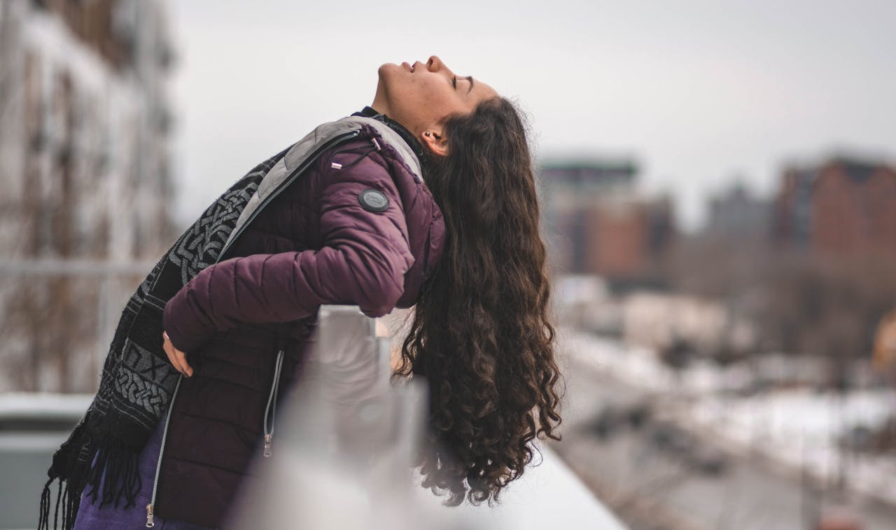 A woman with long hair stands on a balcony, gazing out at the view with a serene expression.