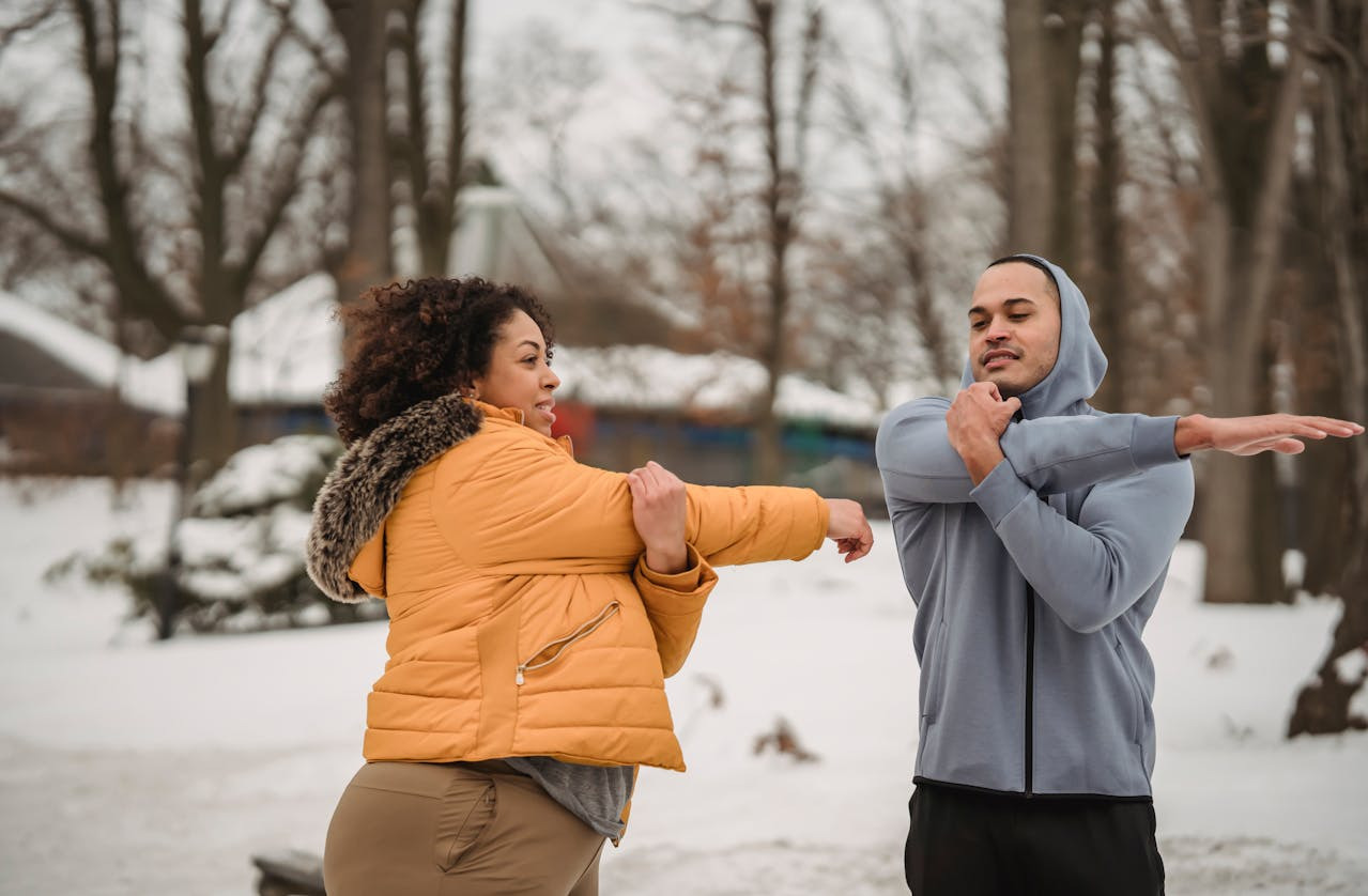 A man and woman stretch in a snowy park. She wears a yellow jacket, he a gray hoodie. Bare trees and snow set a peaceful winter exercise scene.