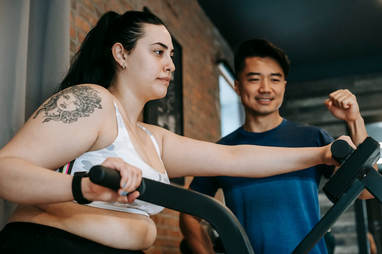 A woman exercising on a stationary bike in a gym, focused and determined. A man beside her, smiling supportively, creates a motivational atmosphere.