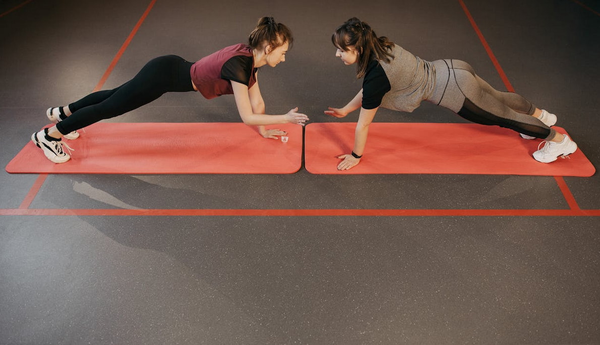 Two women engage in a plank challenge on red exercise mats, facing each other in a gym setting. The mood is focused and cooperative.