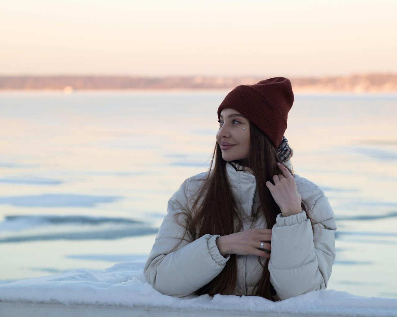 A woman in a white coat and red beanie sits on snowy ground by a frozen lake. She smiles softly, her long hair falling over her shoulders. Serene winter scene.