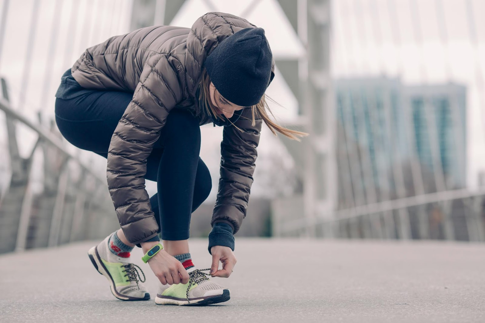 Woman in a winter jacket and beanie crouches to tie her running shoe on a bridge. The scene conveys determination on a chilly day.