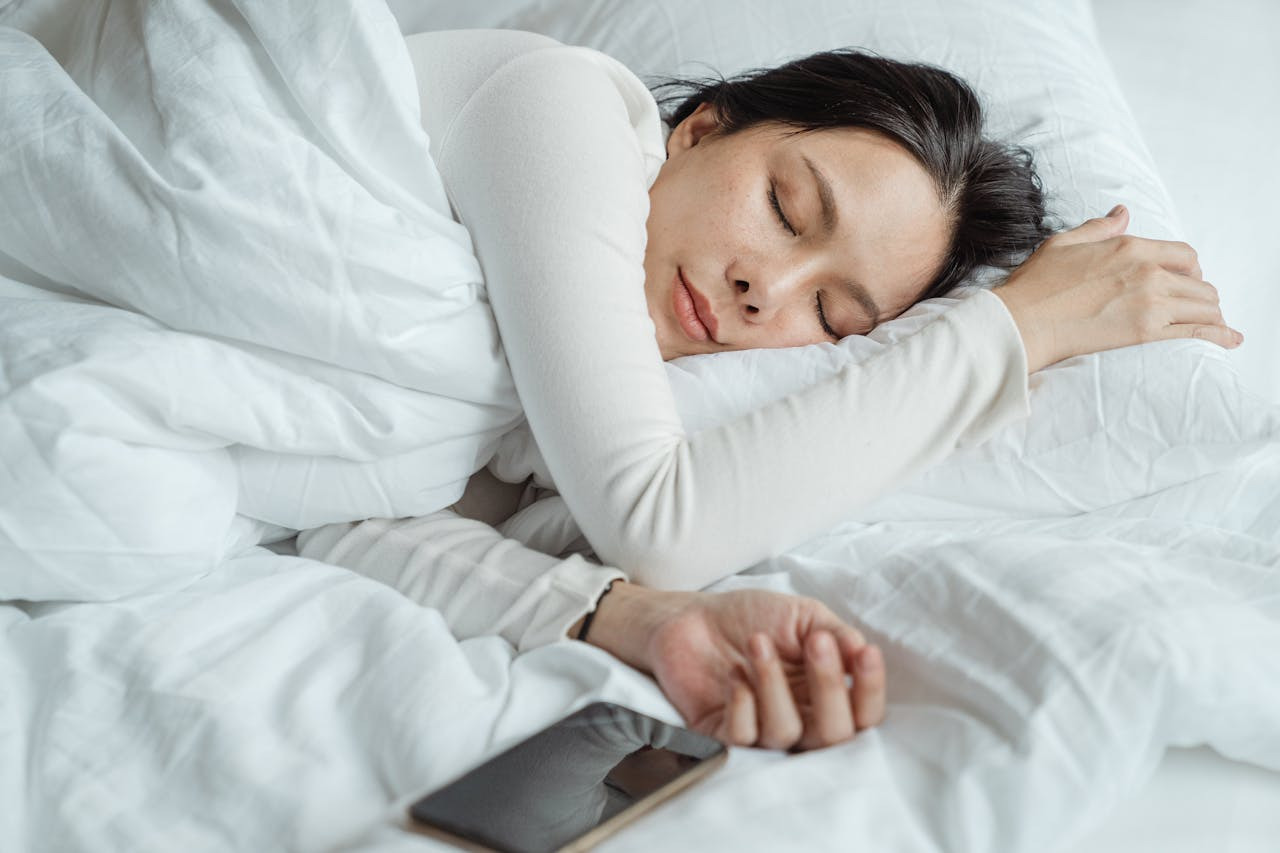 A woman peacefully sleeps on her side, head resting on a white pillow. She wears a white top, with a calm expression in a bright, serene bedroom.