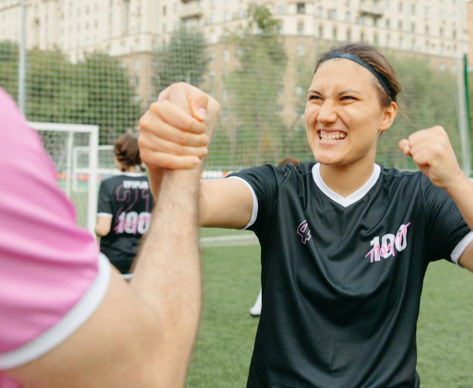 A woman in a black sports jersey, smiling with determination, clasps hands firmly with a teammate. A soccer field and net are in the background.