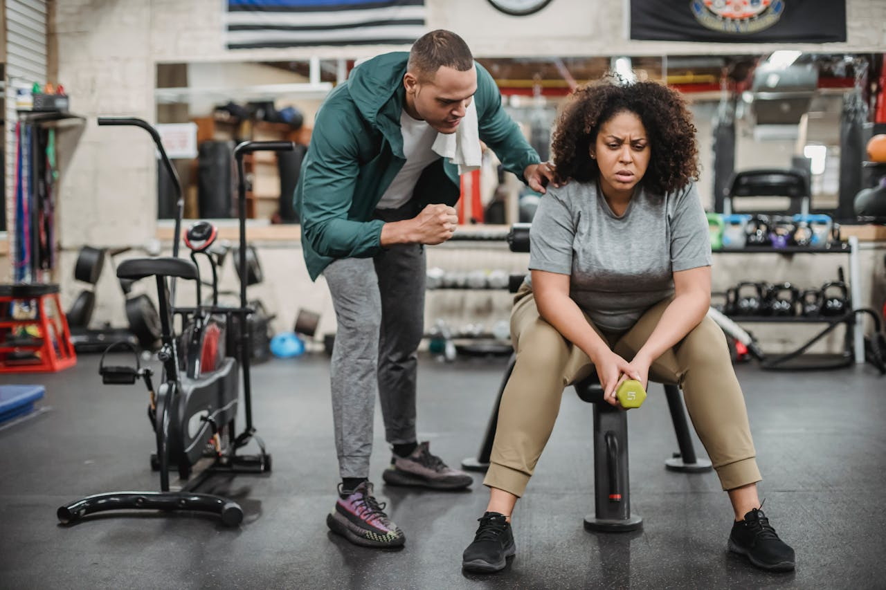 A person in a gray shirt holds a dumbbell, looking determined and tired on a gym bench. A coach in a green jacket energetically encourages them.