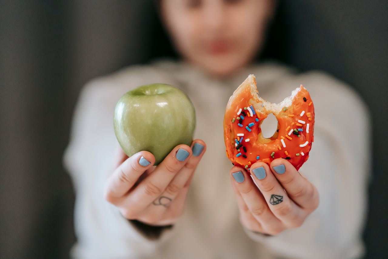 A person with blue nail polish holds a green apple in one hand and a bitten, sprinkle-covered donut in the other, symbolizing a choice between healthy and indulgent foods.