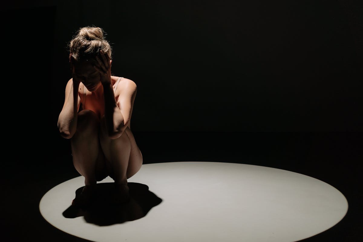 woman curled in a posture of anxiety, sitting in a spotlight in an otherwise dark frame