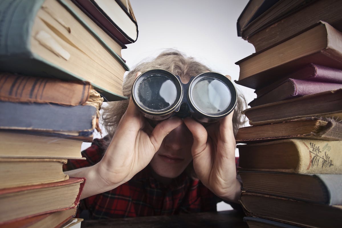 person with binoculars peeking between two towering stacks of books to indicate searching for mental clarity and foucs