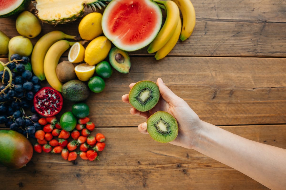 woman holding sliced kiwi in front of a table strewn with other healthy fruits