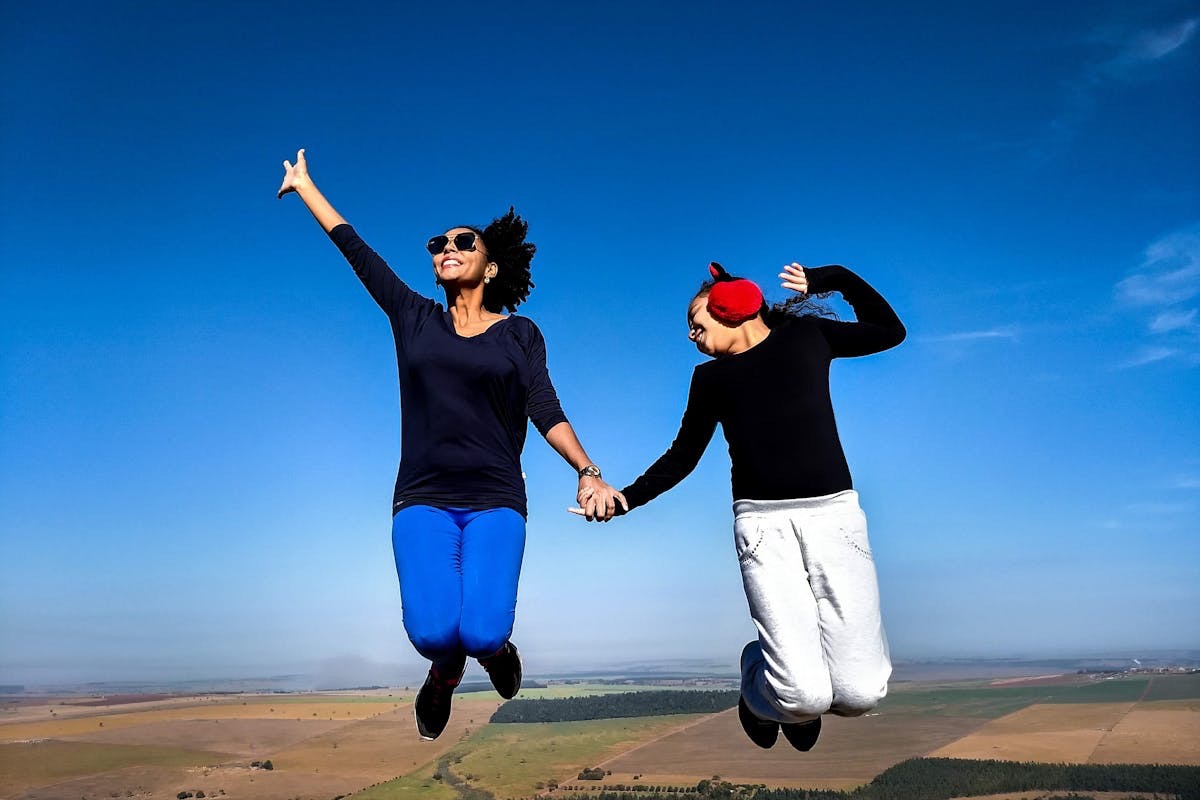 two women holding hands and jumping in an expression of joy against a bright blue cloudless sky 