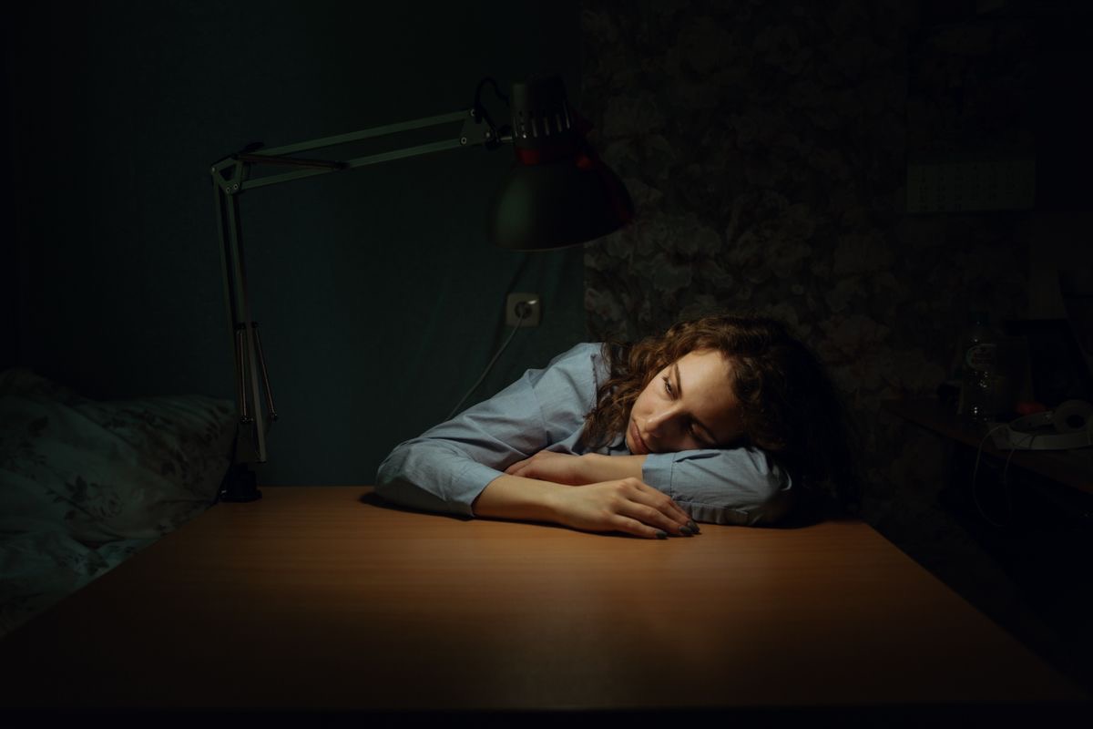 woman looking exhausted as she rests her head on a desk in the glow of a table lamp