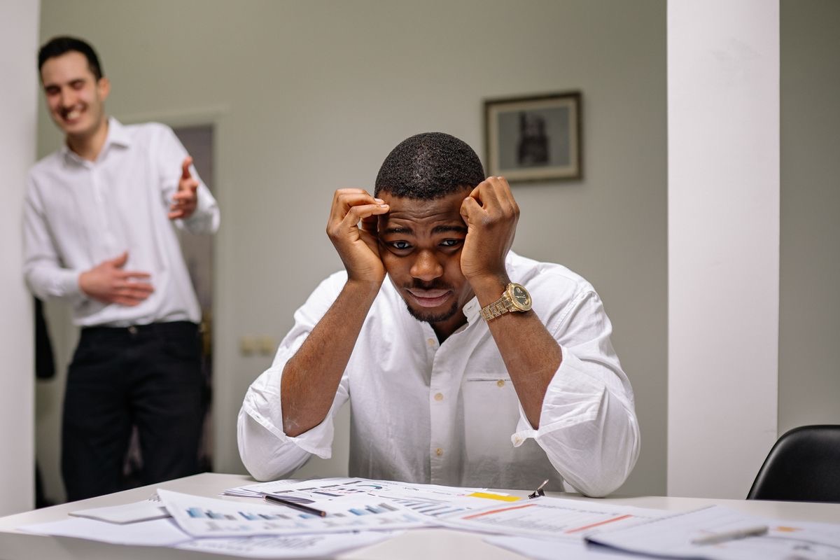 man stressing out at a table as another man laughs at him in the background