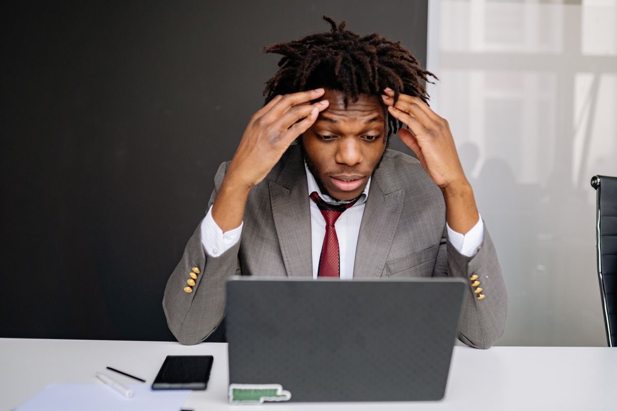 man in a suit holding his forehead, looking at his laptop with a stressed out expression