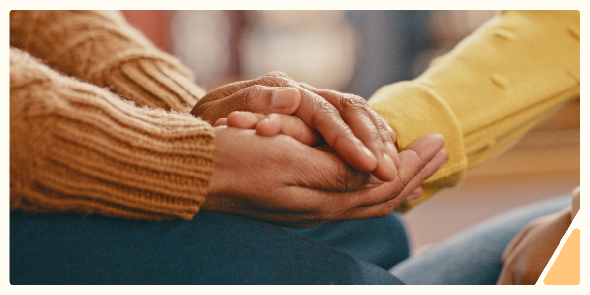 Two people holding hands in comfort, framed with soft edges and an orange corner design, representing support for Suicide Prevention Month.