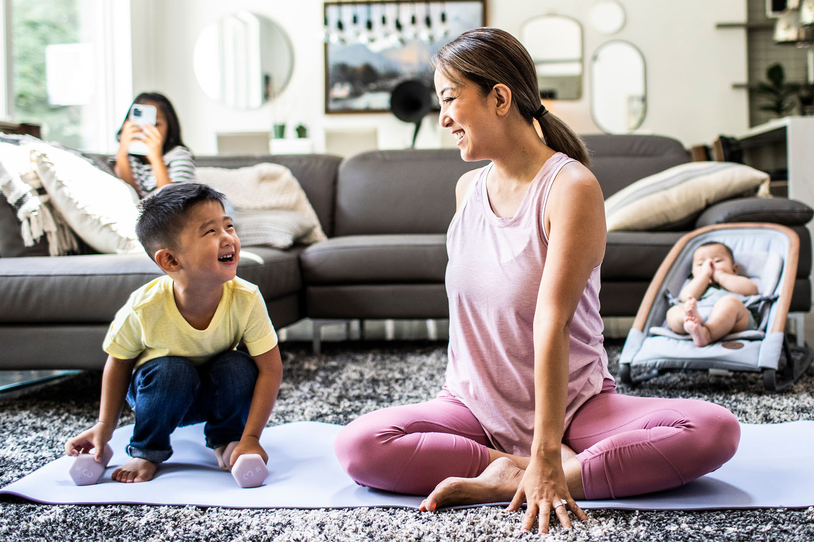 A woman in athletic clothing poses on a workout mat with young children around her in a living room setting.