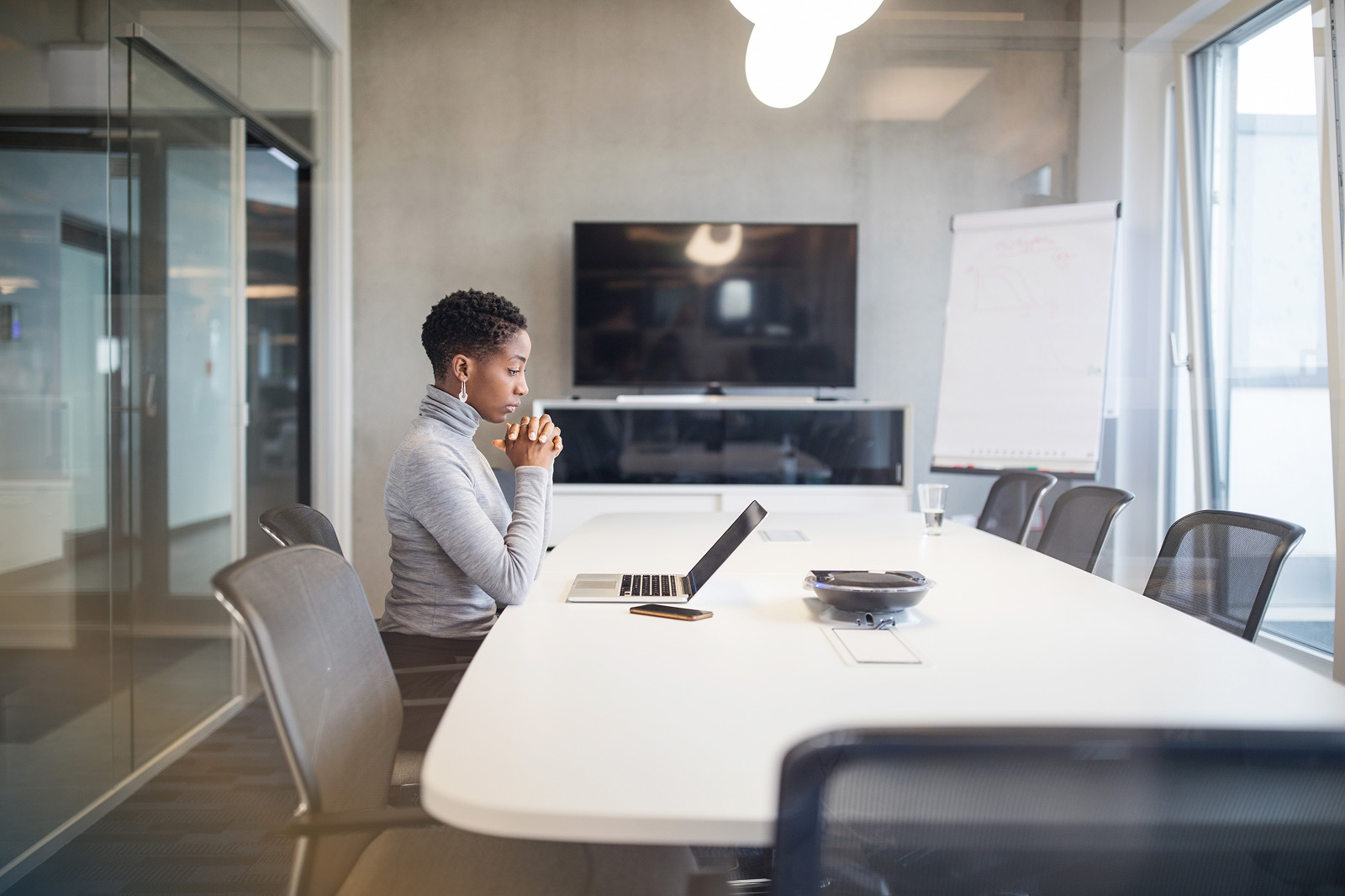 A woman sits alone at a conference room table looking at her laptop with a stressed, focused expression.