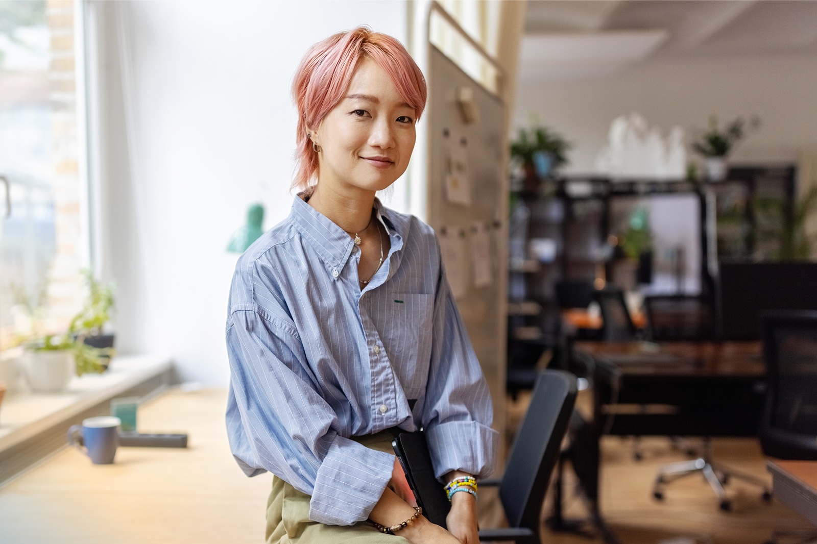 A person with short pink hair sits in a bright, modern office space, smiling gently toward the camera. They are wearing a light blue button-down shirt and holding a notebook or tablet on their lap. Large windows provide natural light, and desks, chairs, an