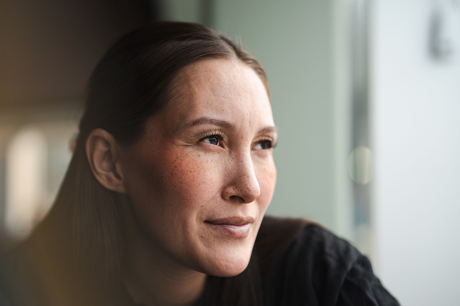 A person with freckles and brown hair gazes thoughtfully out a window, their expression calm and reflective.