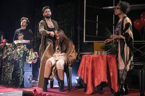 Performers on stage at a red table, Verona Koxha (Cassandra), a white woman, is seated and Ivan Mihailović (Soldier), a white man, is standing and forcing her to sing