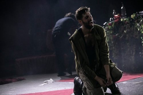 Ivan Mihailović (Solider), a white man, on stage and kneeling on the floor, he is dimly lit