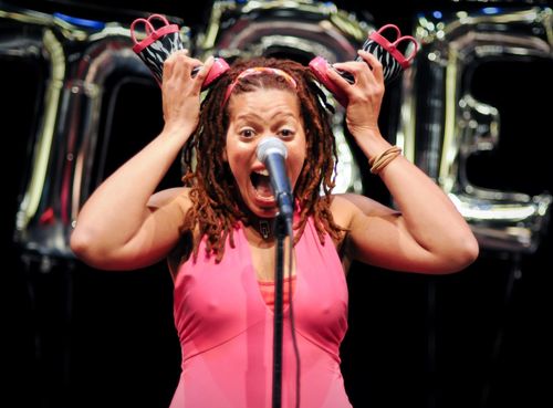 Meredith Wright, a singer with brown braids, is at the microphone with her mouth open, she is wearing a pink dress and holding two pink and zebra print boots to her ears