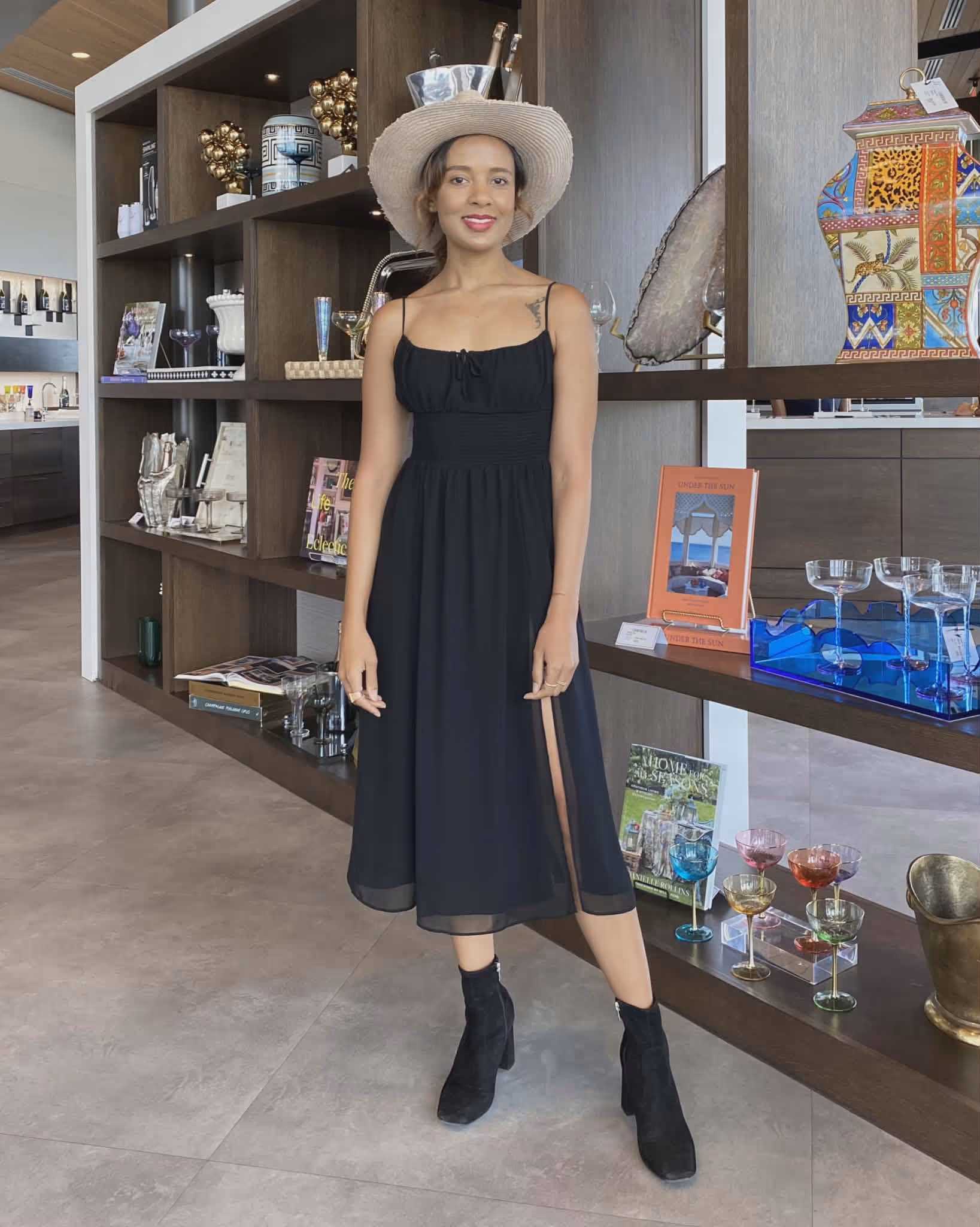 Woman wearing a black sleeveless dress, black boots, and a wide-brimmed hat standing in front of wooden shelves displaying glassware and decorative items.