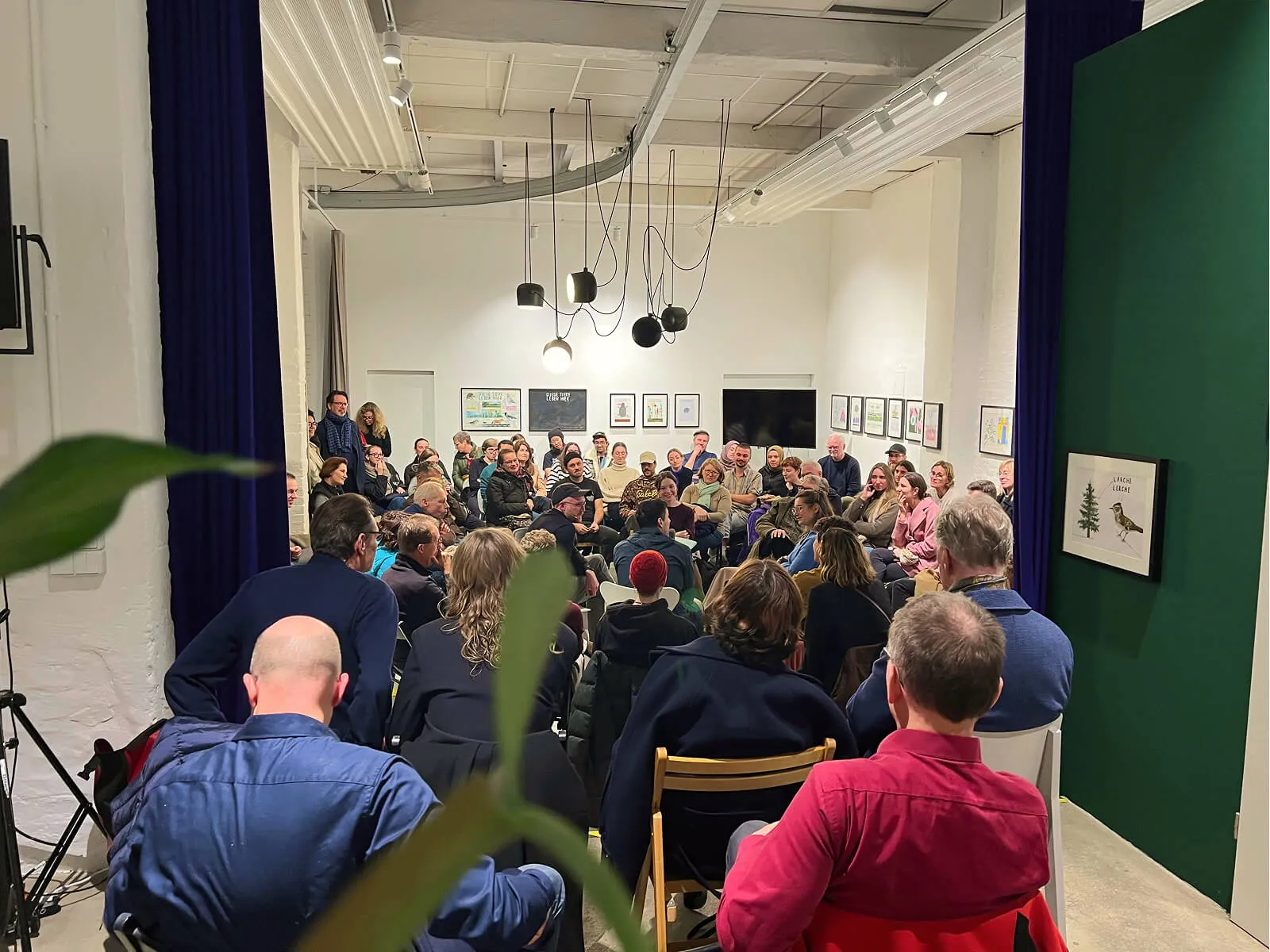 View from behind into the fully occupied event room at GE59 and HENKELHIEDL. The audience sits in a dense circle of chairs and looks towards the center; a plant can be seen blurred in the foreground.