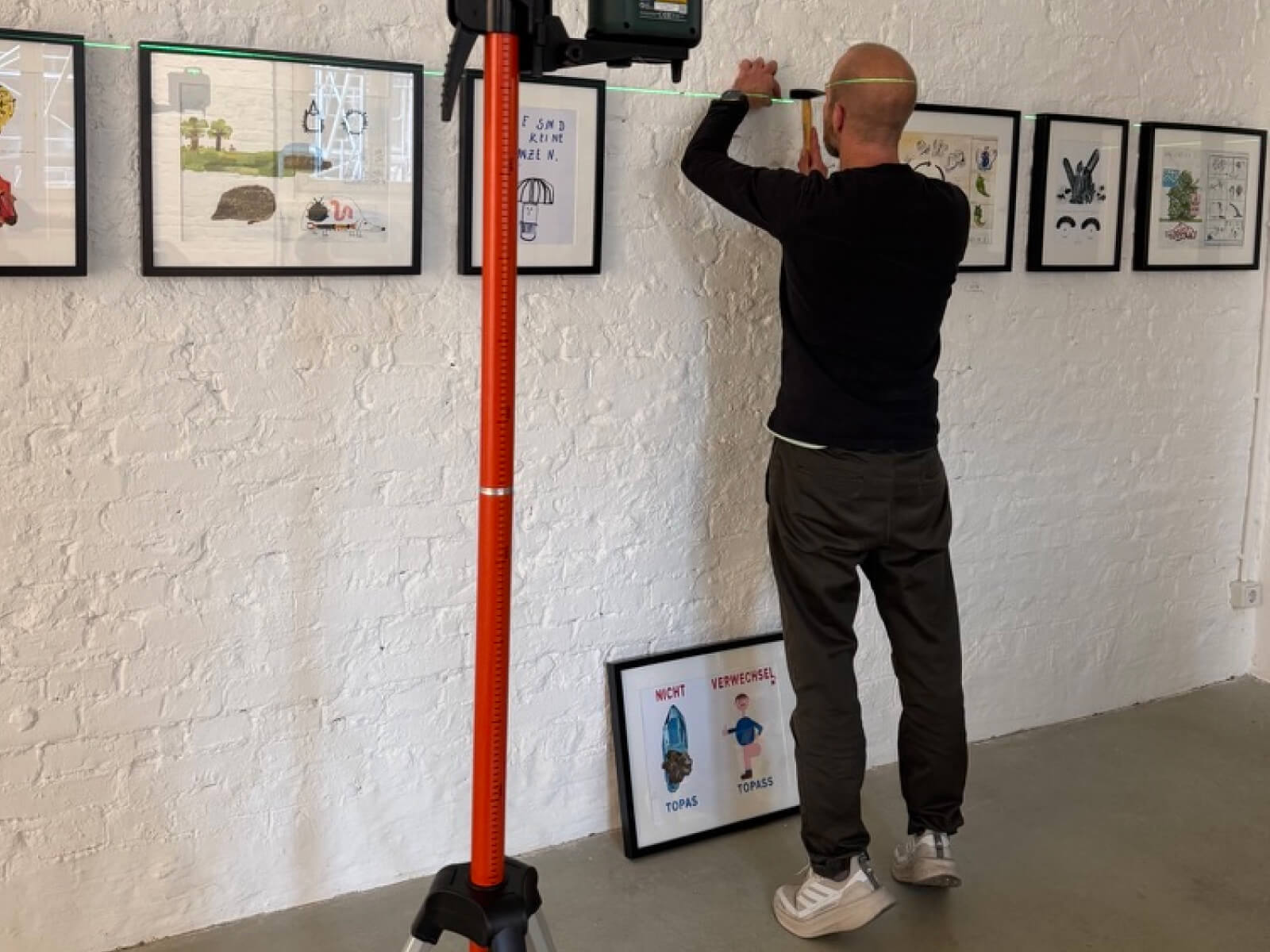 Andreas Henkel hängt mit einem Hammer und einer Laser-Wasserwaage ein gerahmtes Bild von Felix Bork an eine weiße Ziegelwand für die SIMPLEX-Ausstellung.