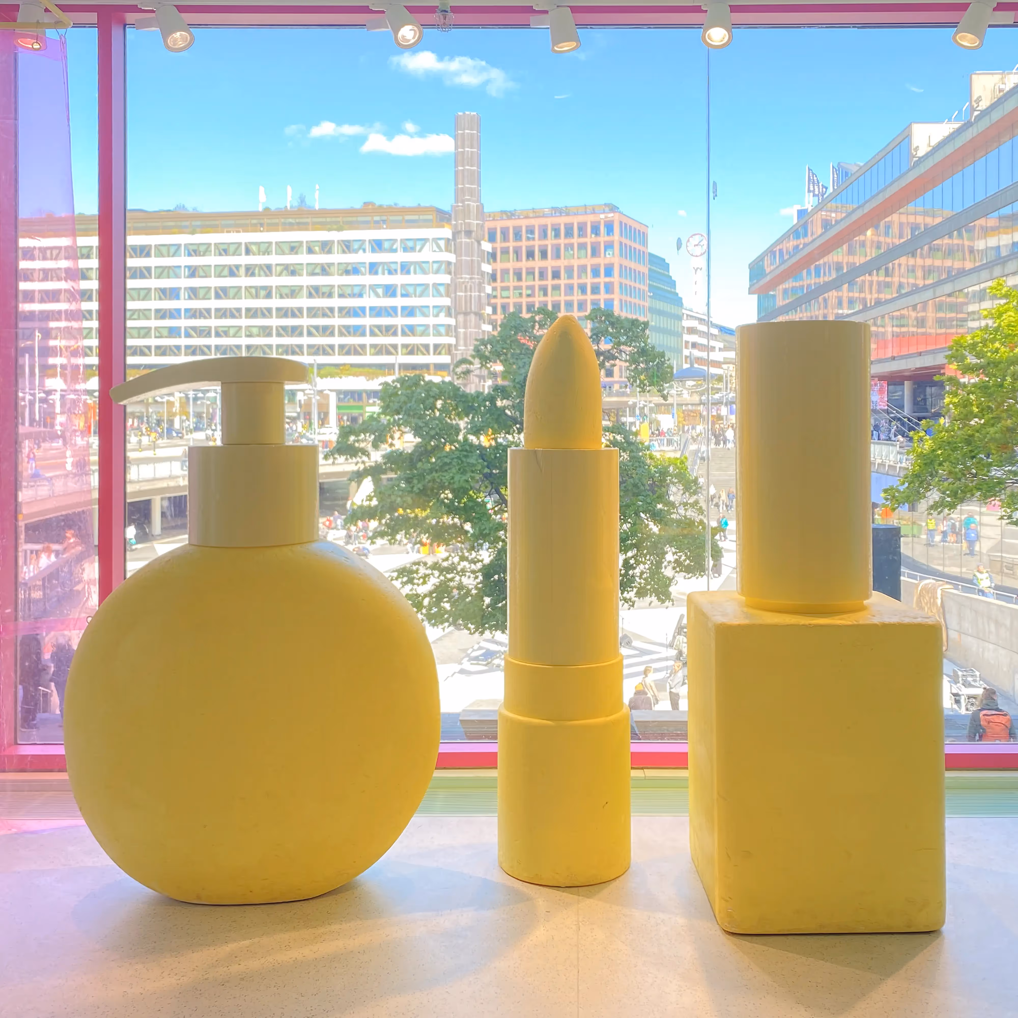 Yellow sculptures of beauty products set against a city view through pink-tinted windows