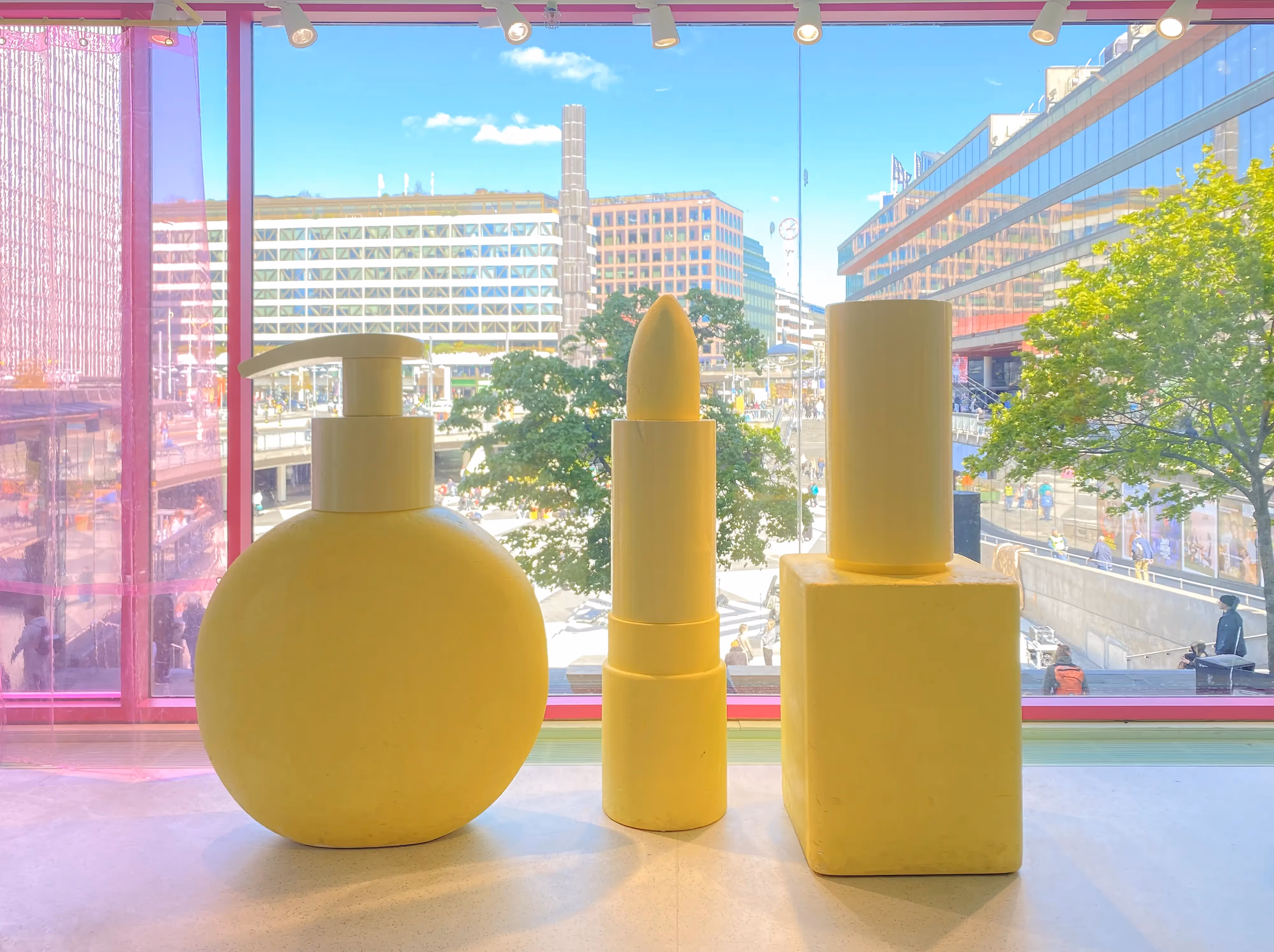 Yellow sculptures of beauty products set against a city view through pink-tinted windows