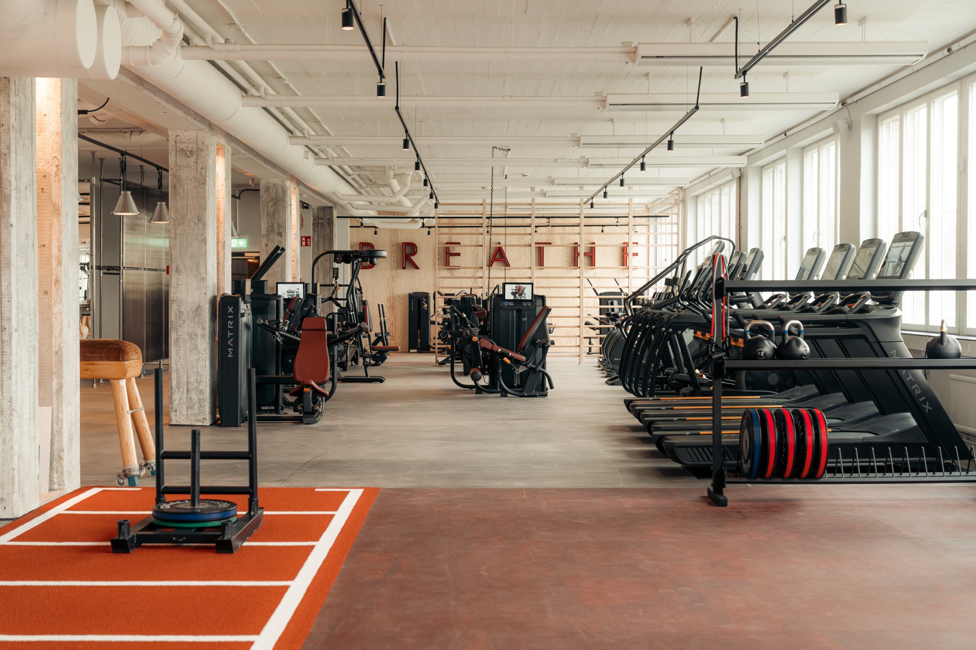 Daylit gym with concrete columns, a red training lane and BREATHE lettering at Mornington Health Club Bromma.