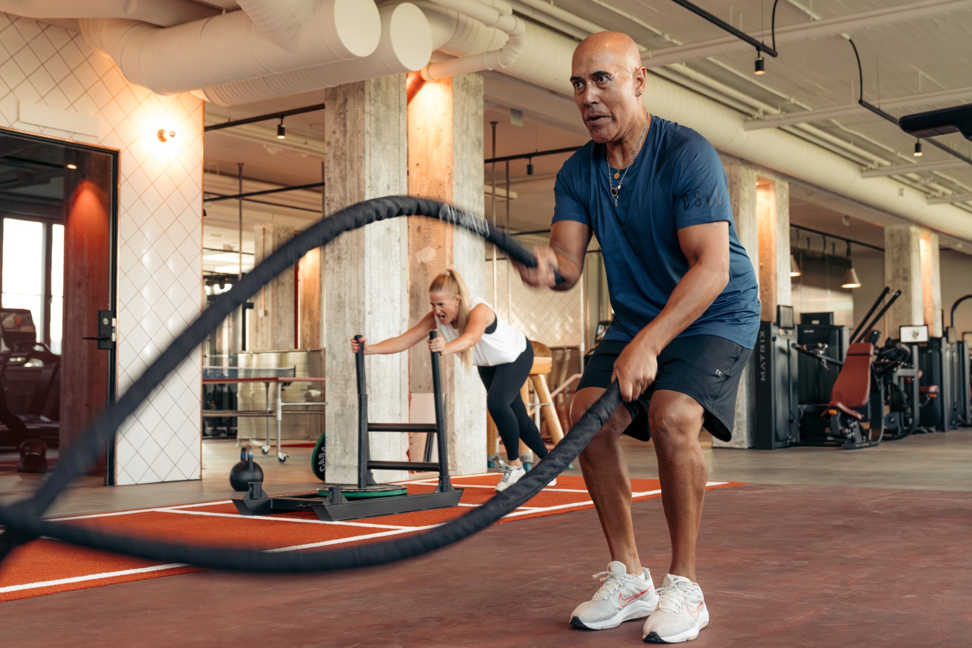 A member uses battle ropes on the red training floor beside exposed concrete columns.