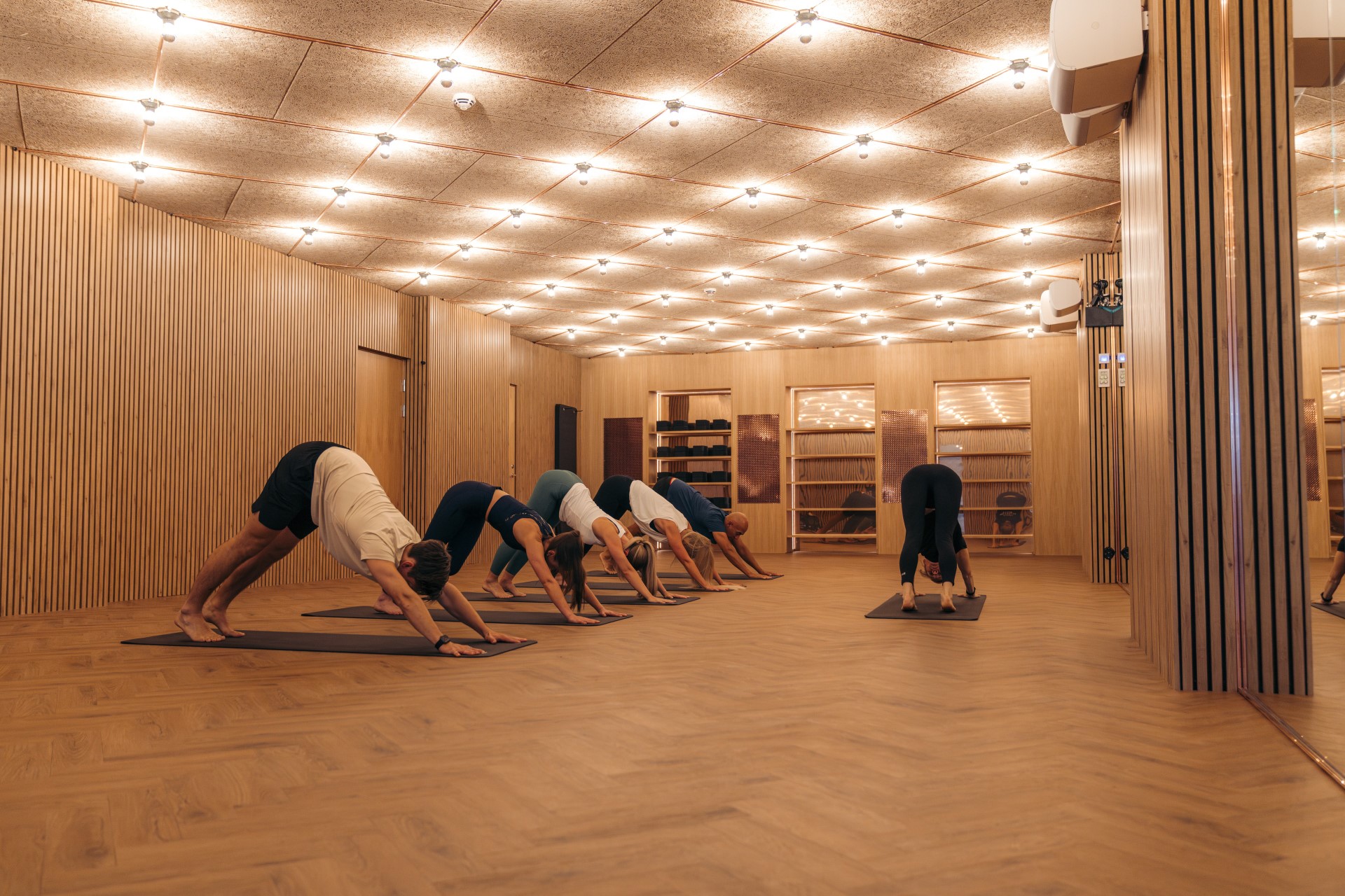 Group stretching in a timber-lined studio with herringbone flooring and warm ceiling lights.